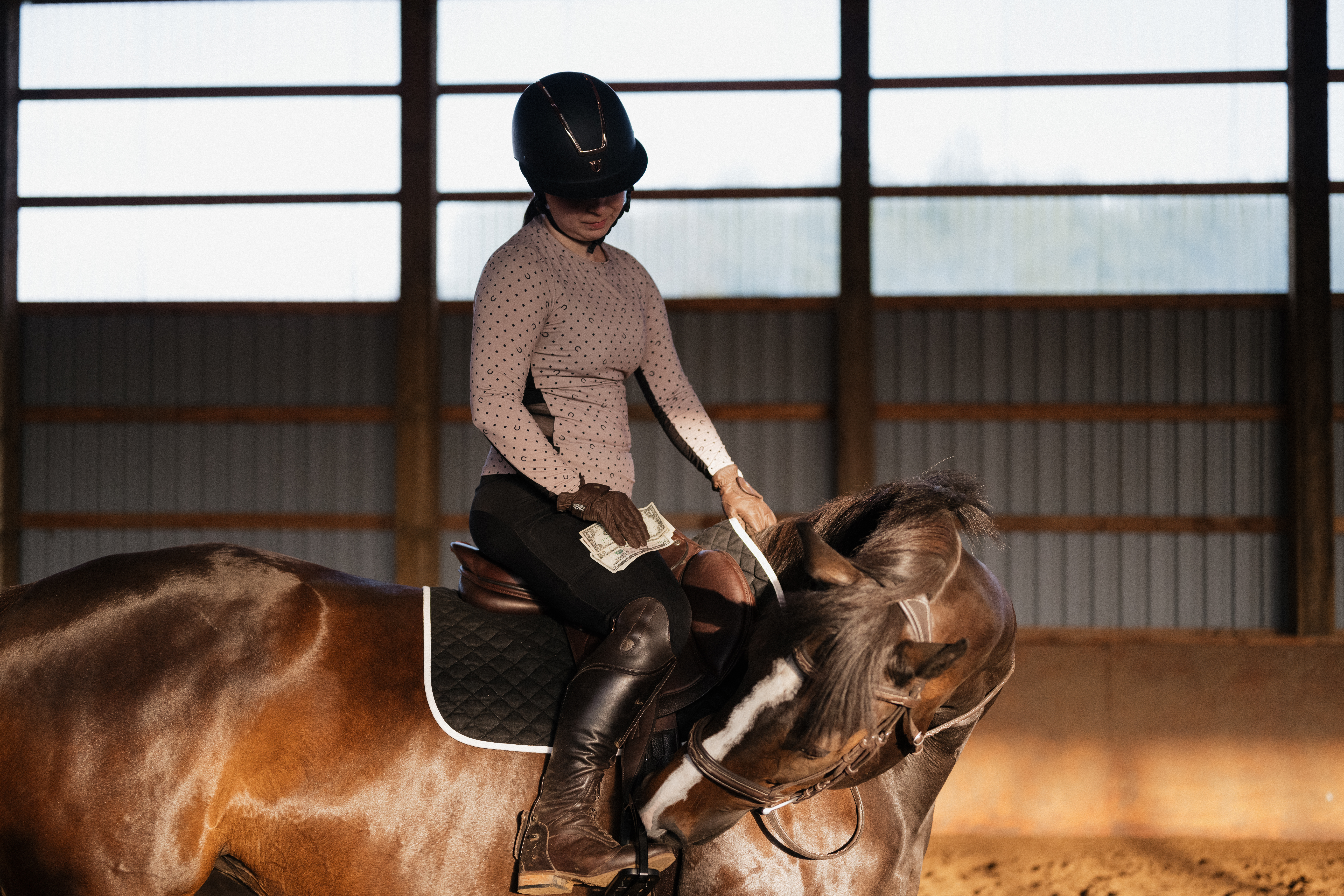 A rider in a helmet and equestrian gear sits atop a brown horse in an indoor arena, holding cash in one hand while guiding the horse with the other. The natural light filters through the windows, highlighting the connection between the rider and horse, symbolizing financial considerations in the equestrian world