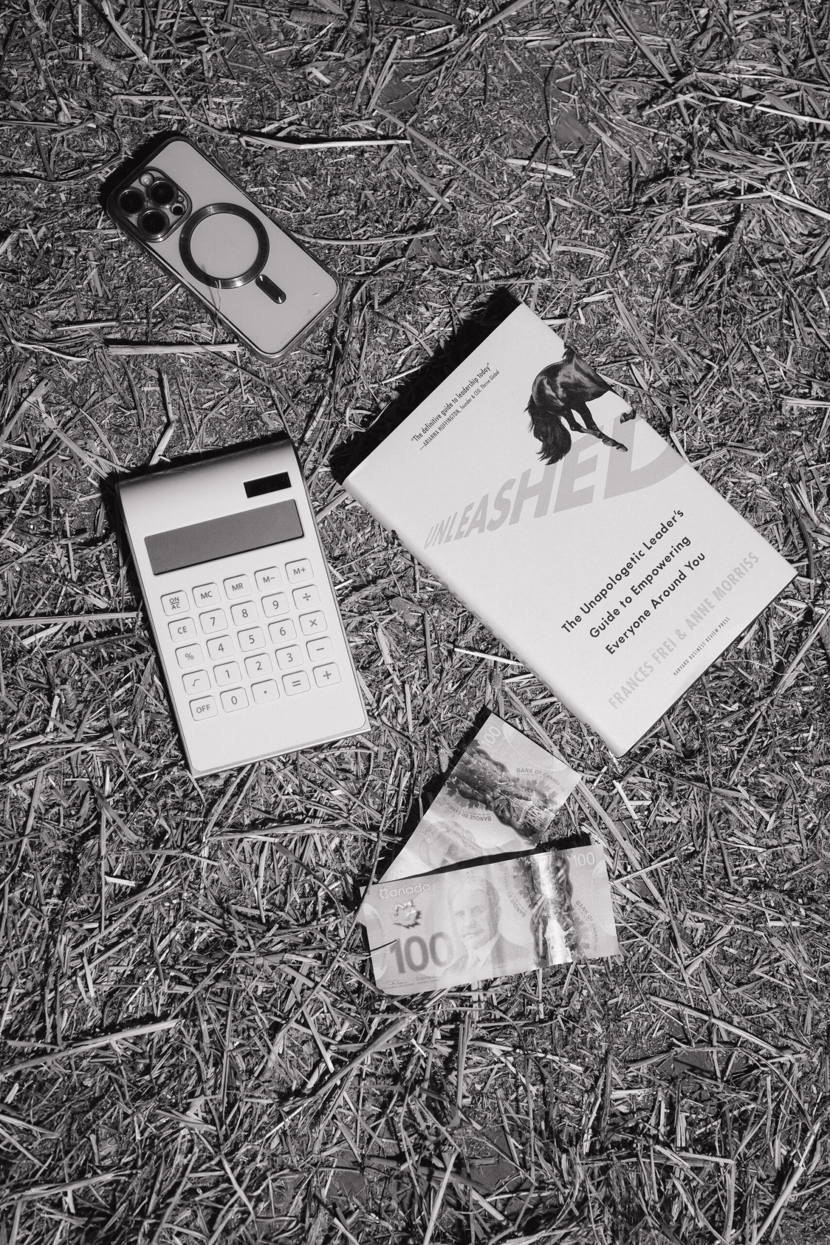 Black and white photo of a flat lay on hay featuring an iPhone, a white calculator, two Canadian $100 bills, and a book titled Unleashed: The Unapologetic Leader’s Guide to Empowering Everyone Around You by Frances Frei and Anne Morriss. The objects are arranged naturally, blending elements of business, money, and leadership within an equestrian setting.